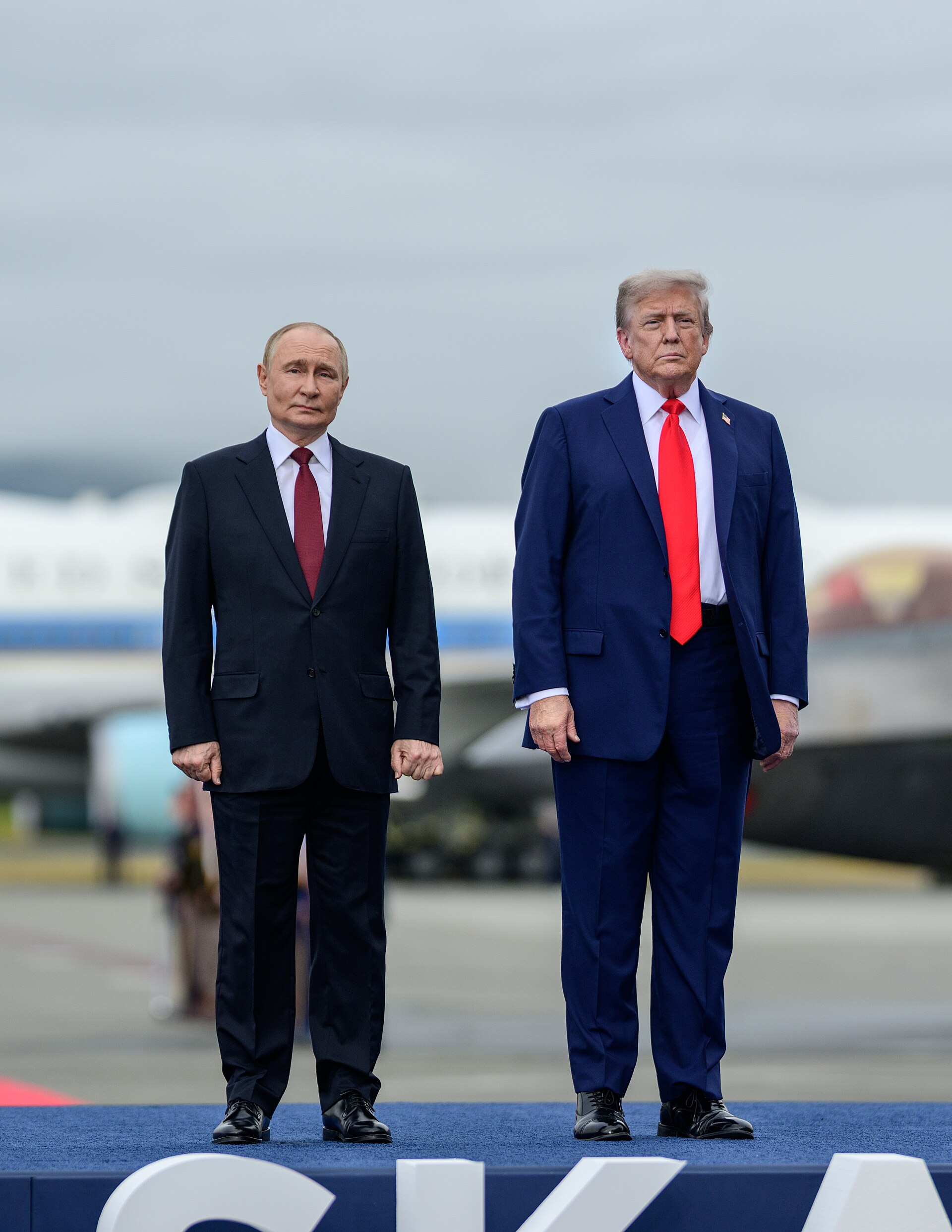 Dos hombres con traje posan de pie, uno junto al otro, sobre una plataforma azul.