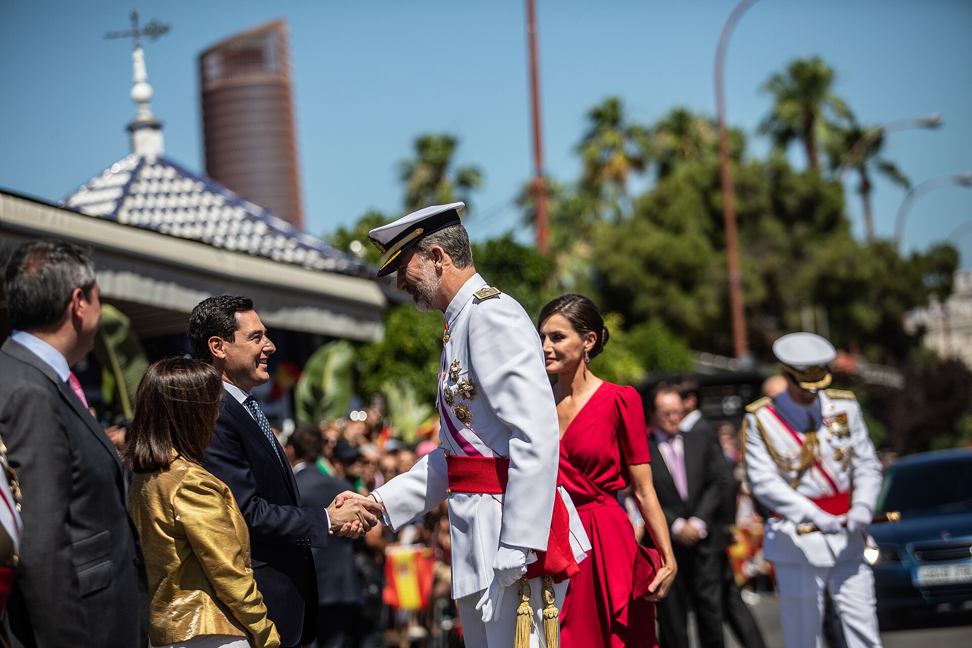 Un hombre con uniforme blanco saluda a otro hombre mientras una mujer con vestido rojo camina detrás; varias personas observan al fondo.