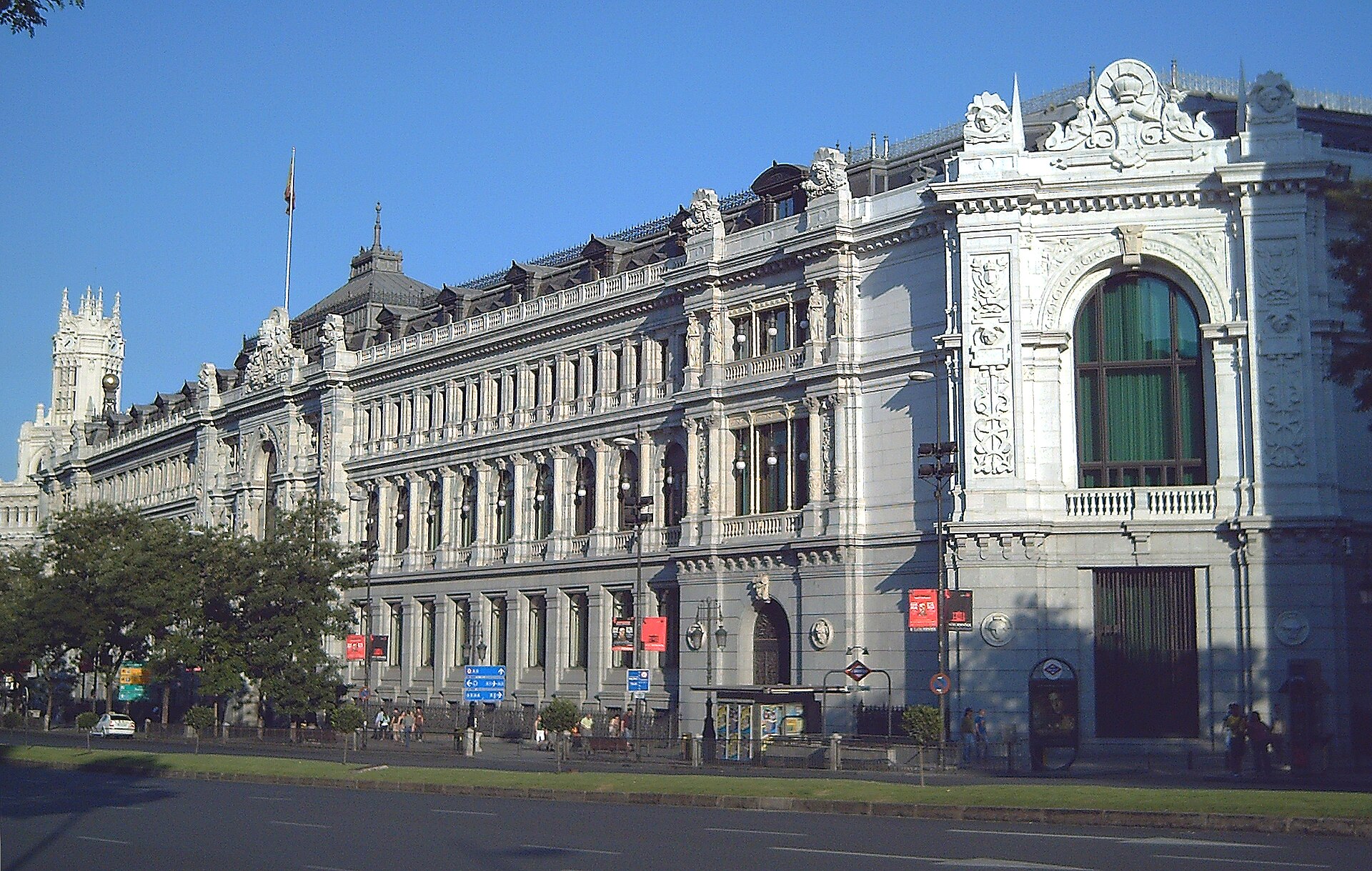 Fachada ornamentada de un edificio institucional, con una bandera y una avenida en primer plano.