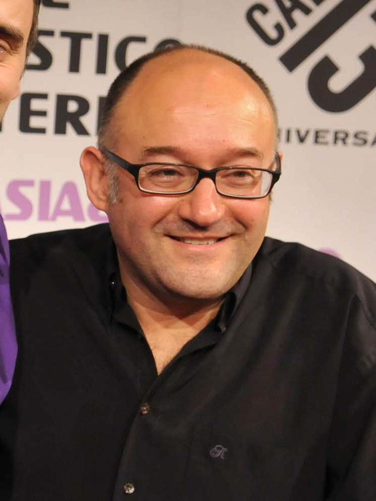 Retrato de un hombre con gafas y camisa negra ante un panel de prensa, junto a otra persona parcialmente visible.