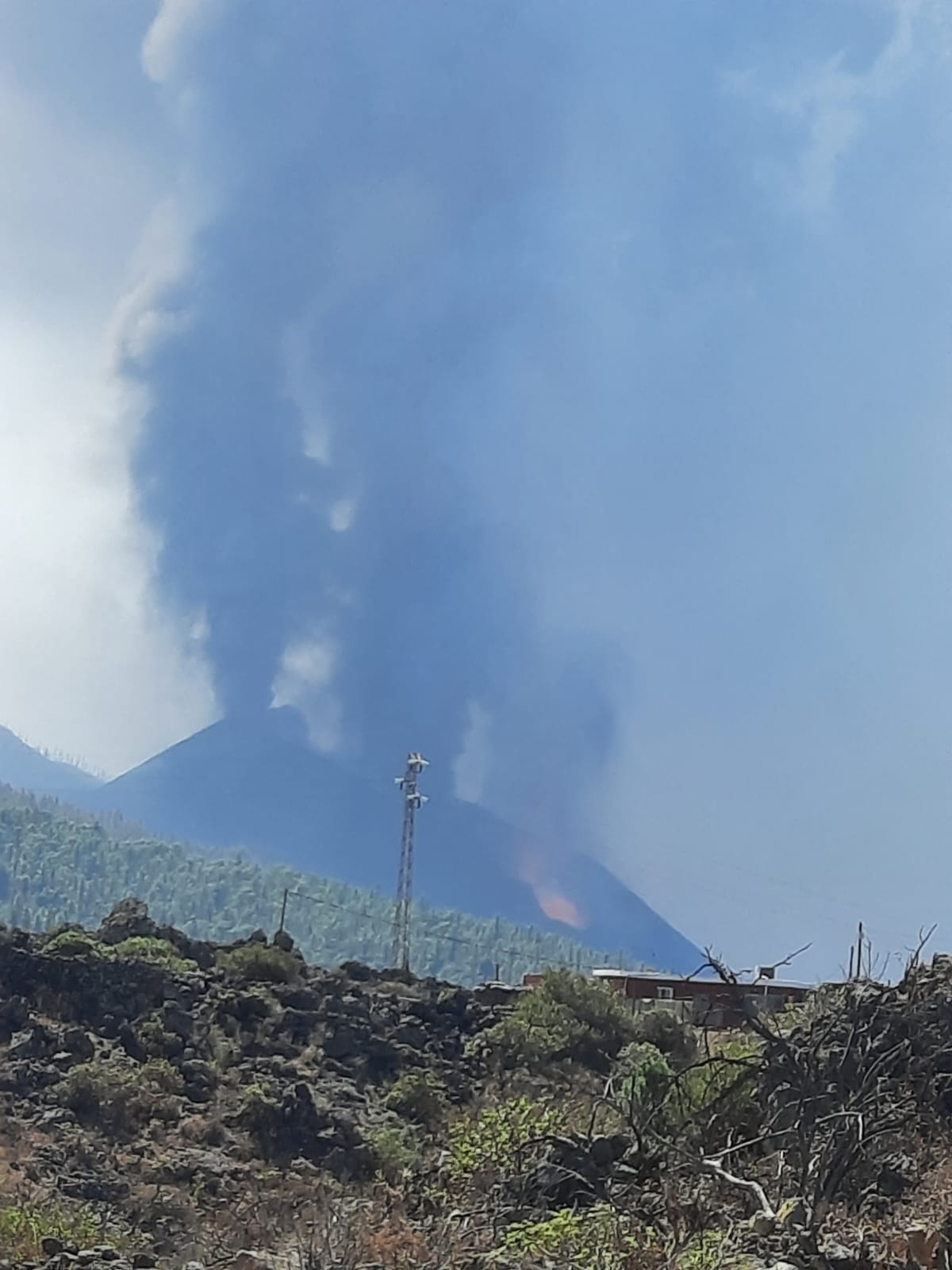 Una columna de humo se eleva tras un terreno cubierto de rocas oscuras, con edificaciones y una torre en primer plano.