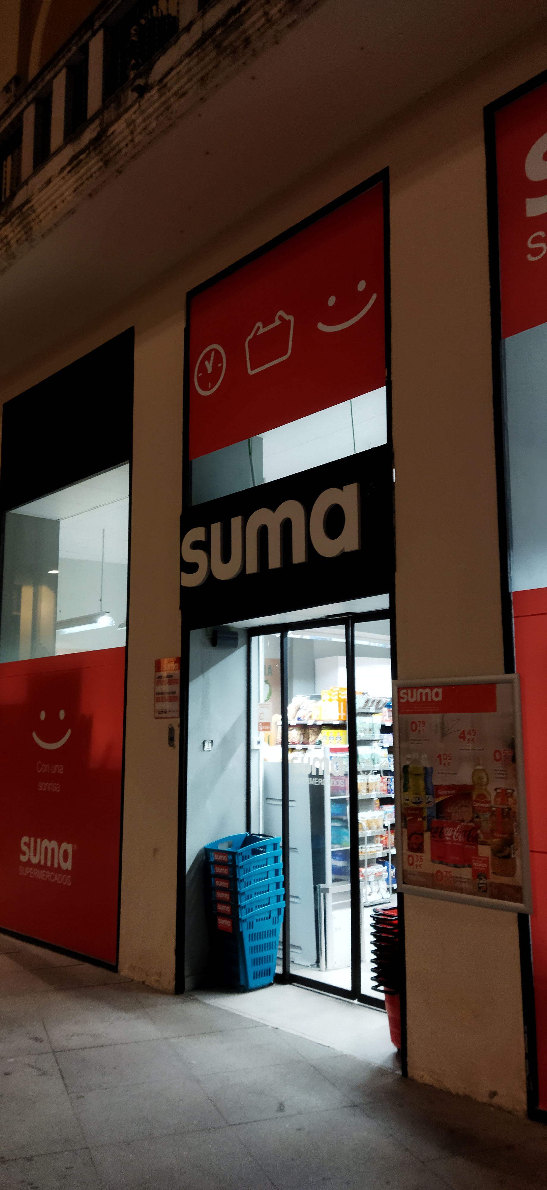 Entrada de un supermercado con cestas apiladas y un cartel de ofertas junto a la puerta.