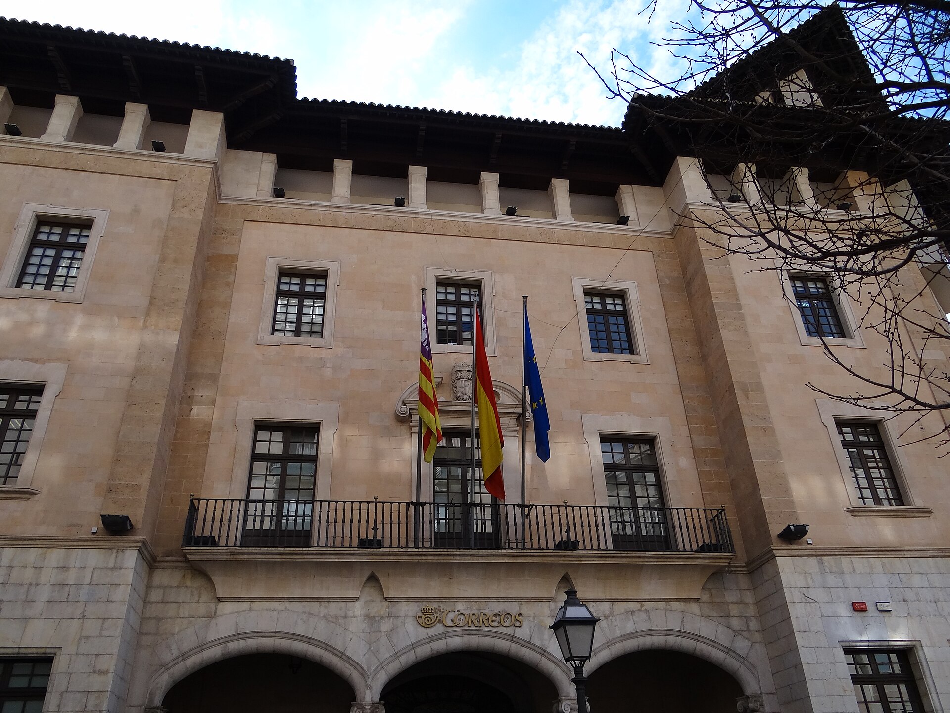 Fachada de piedra con banderas y un rótulo de Correos sobre los arcos.