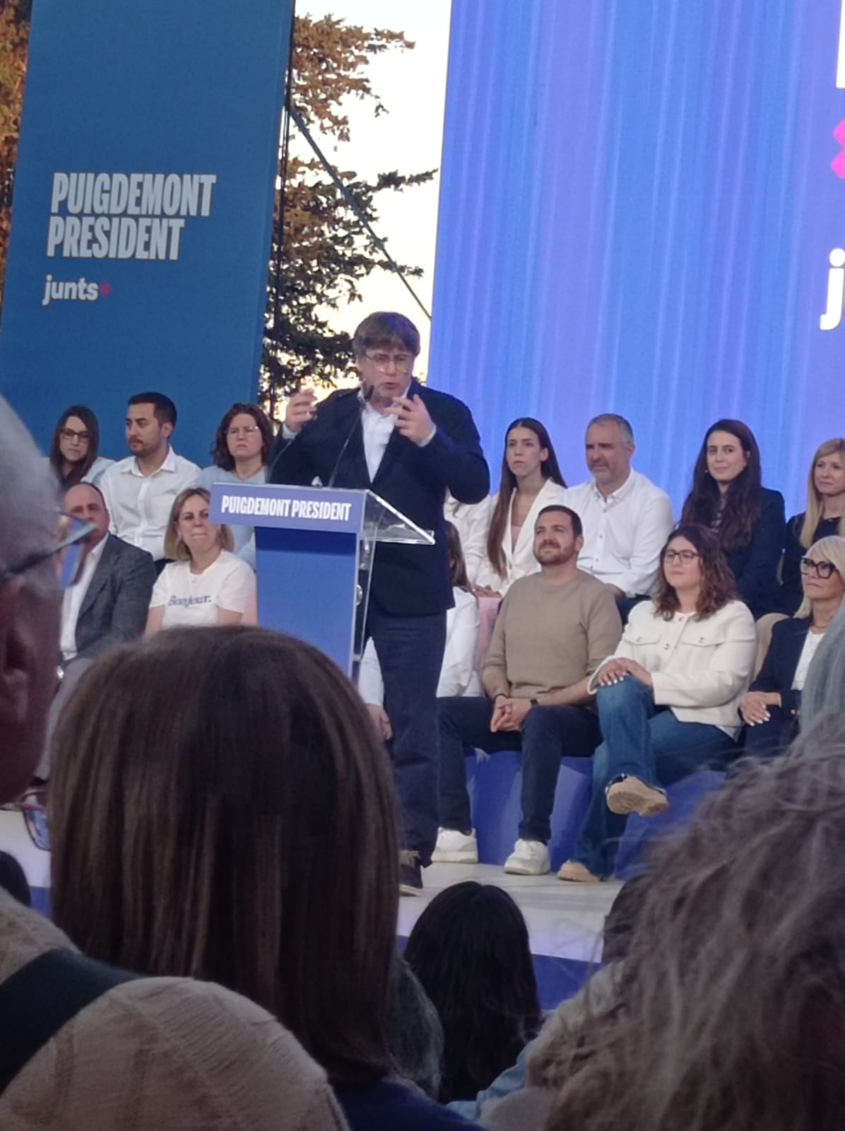 Un hombre habla desde un atril ante un cartel de Junts, con varias personas sentadas detrás.