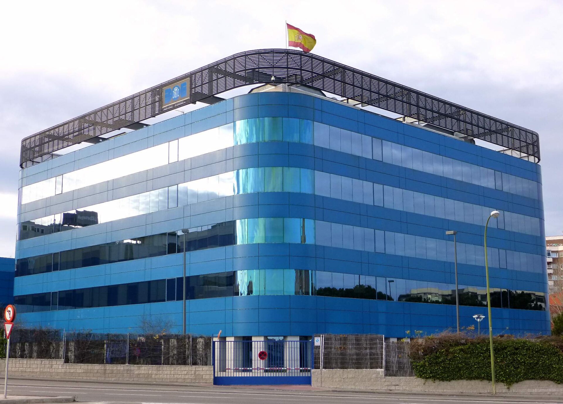 Fachada acristalada azul con una bandera española y un rótulo visible.