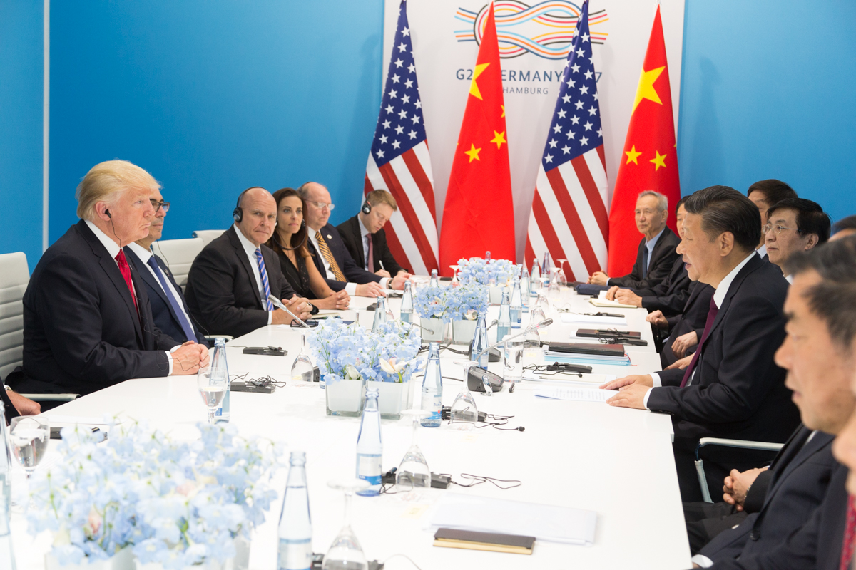 Dos hombres se sientan frente a frente en una mesa de reunión, con banderas de Estados Unidos y China al fondo.