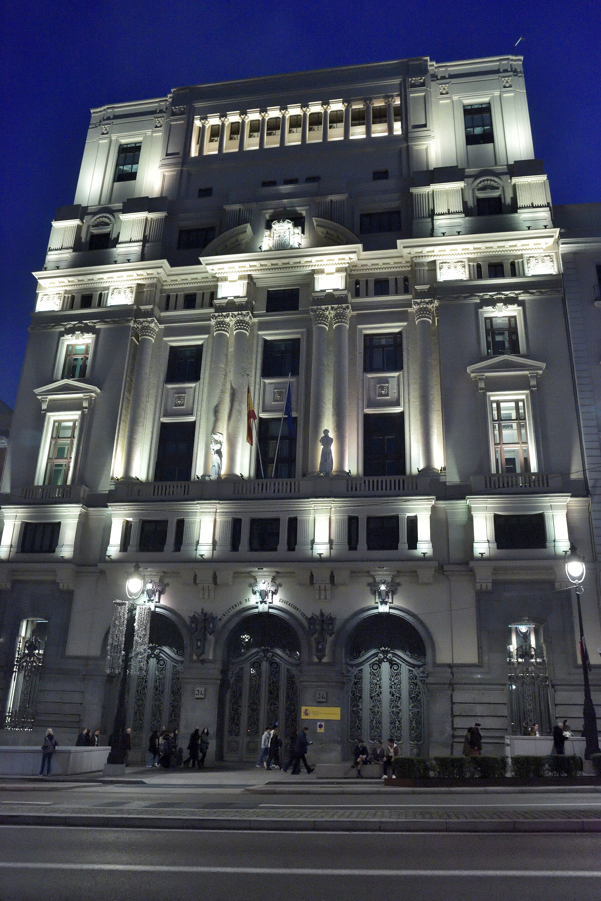 Fachada iluminada de un edificio oficial con el rótulo «Ministerio de Educación y Ciencia» y varias banderas en la entrada.