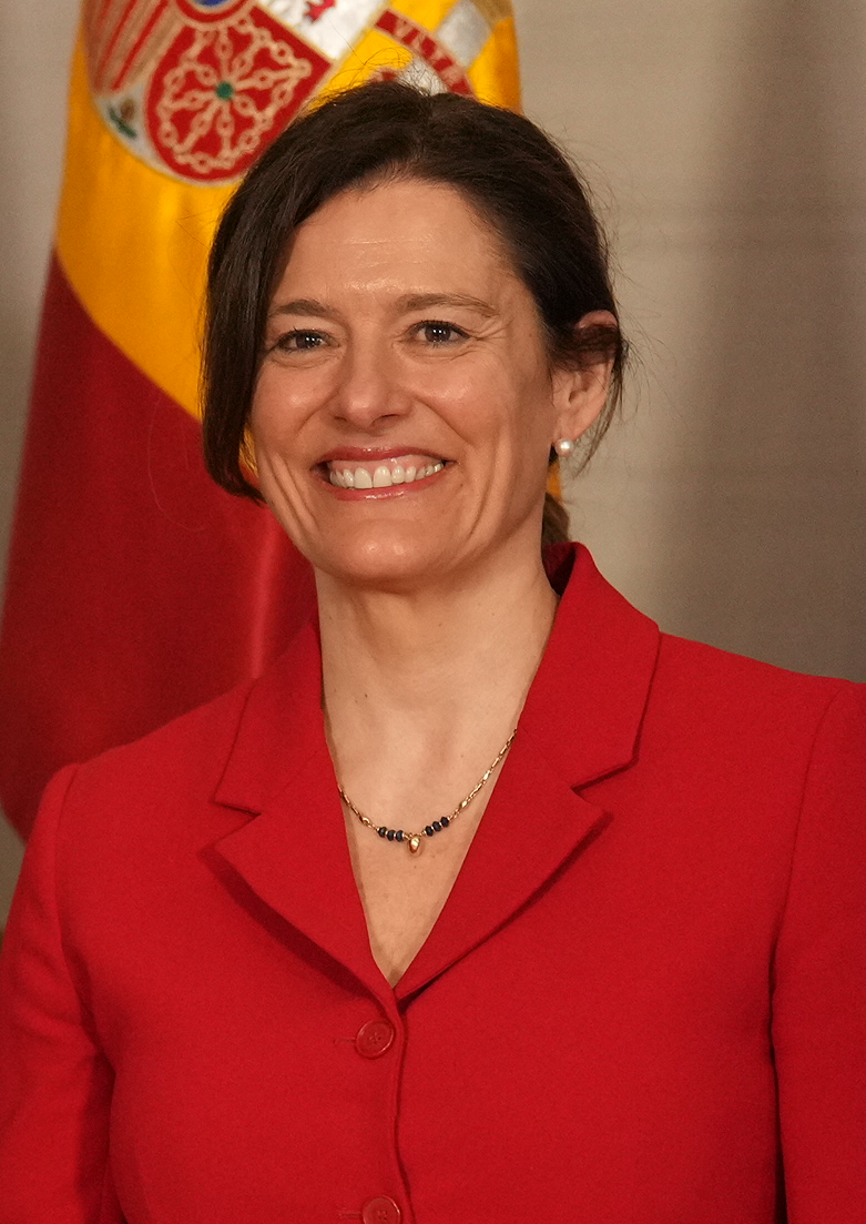 Retrato de una mujer con chaqueta roja, con parte de una bandera española al fondo.