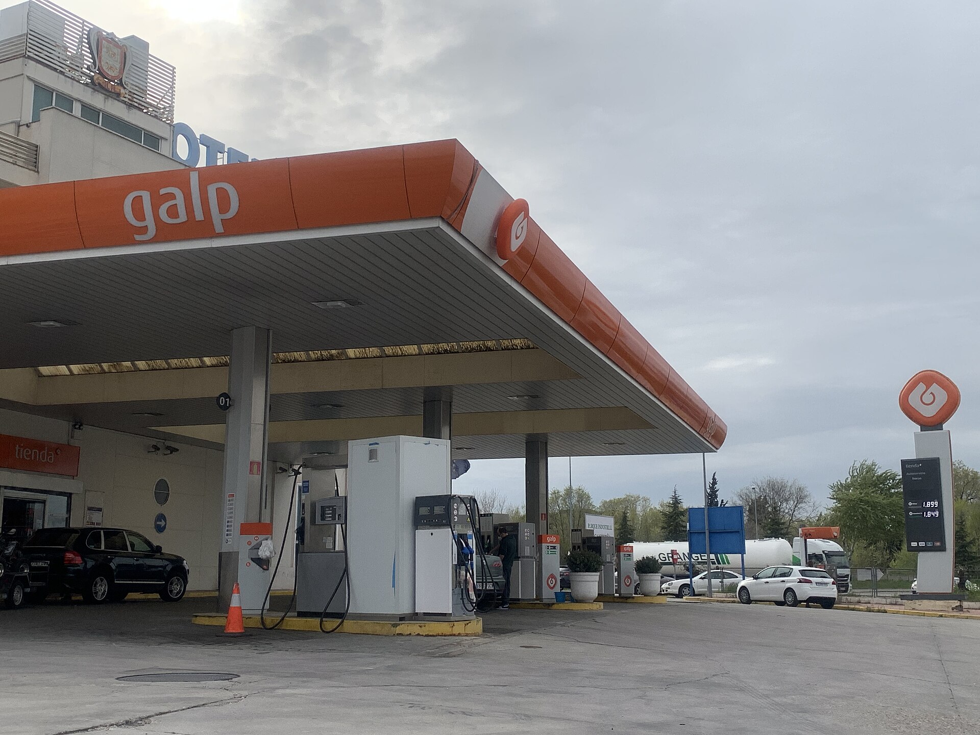 Surtidores de una estación de servicio, con vehículos y un panel de precios al fondo.