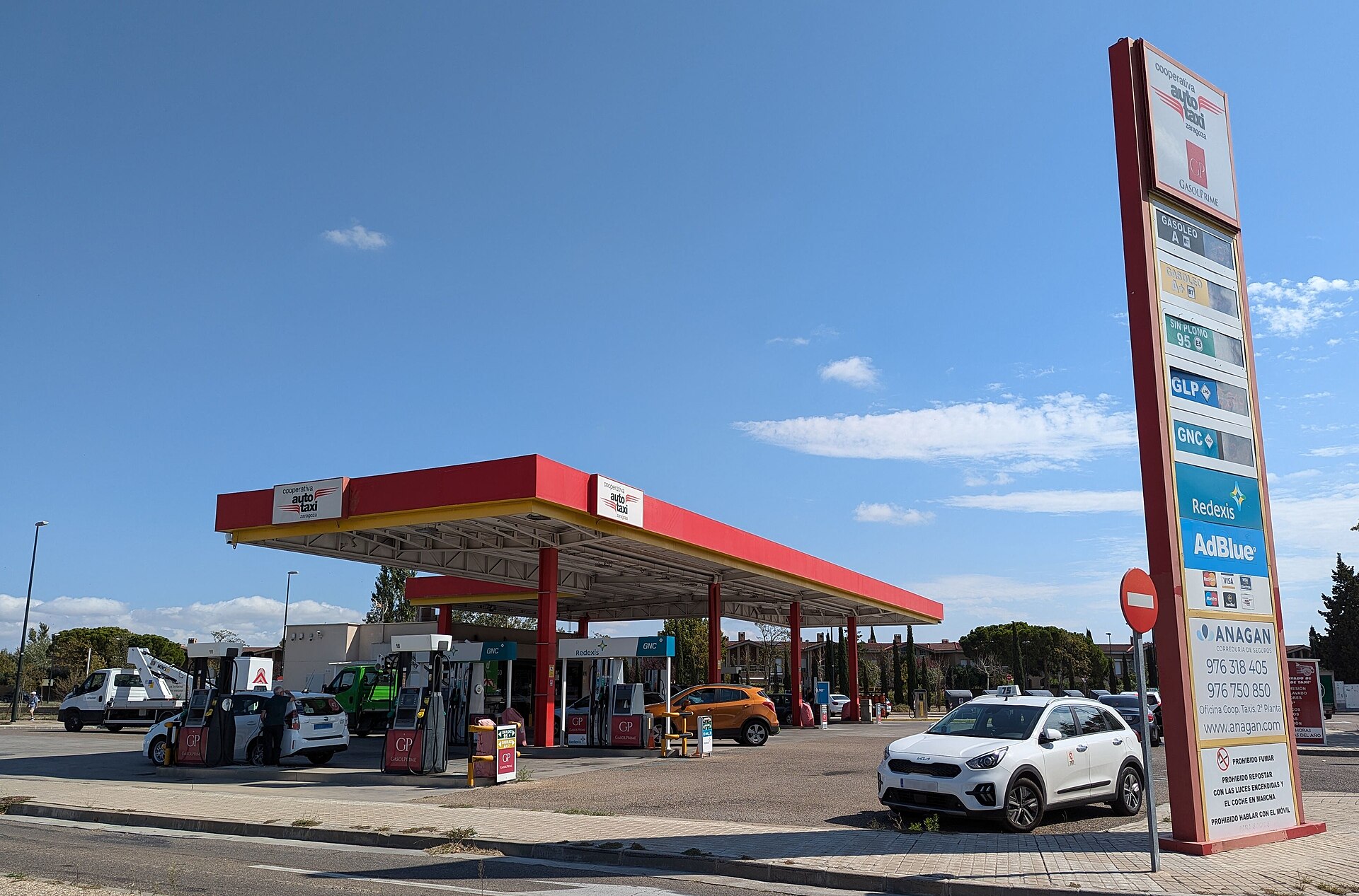 Gasolinera con surtidores, panel de precios y varios vehículos junto a la zona de repostaje.