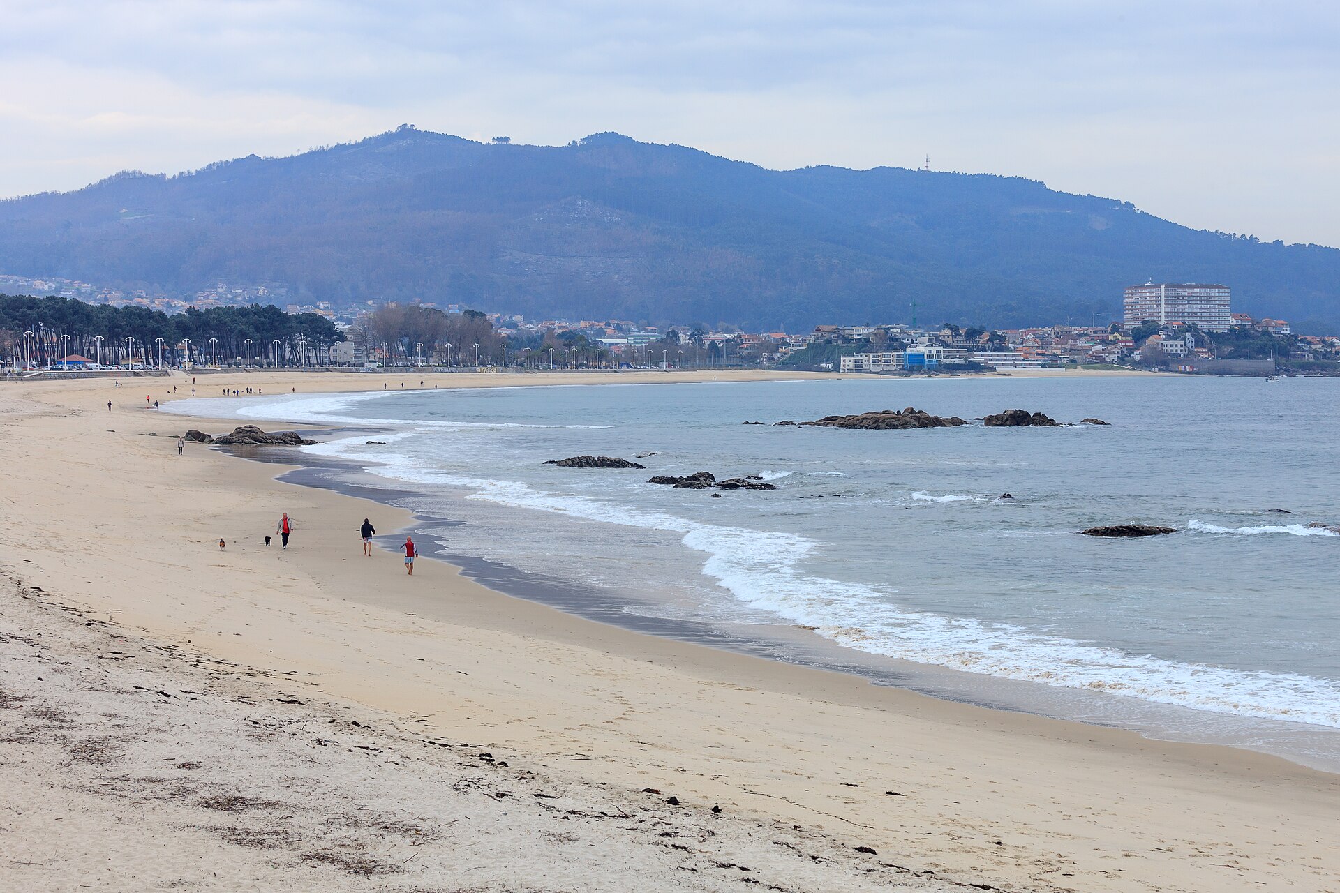 Varias personas caminan por una playa junto a un mar en calma.
