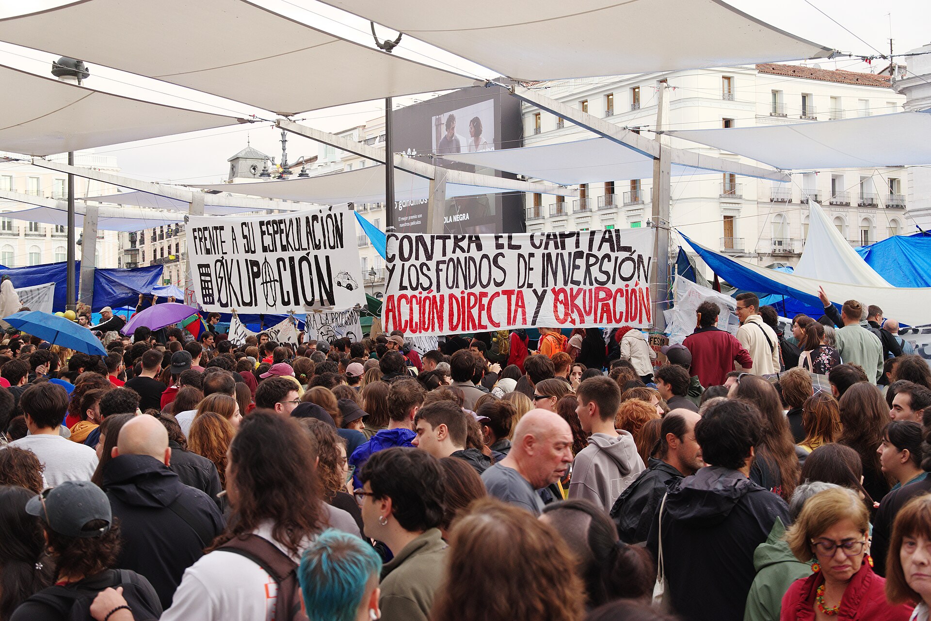 Una multitud se reúne bajo toldos y sostiene pancartas.