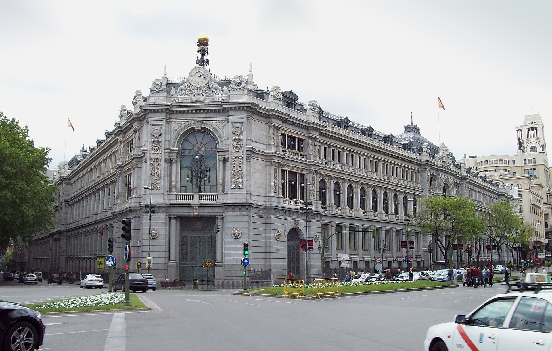 Fachada monumental con un reloj y varias banderas; circulan vehículos en primer plano.