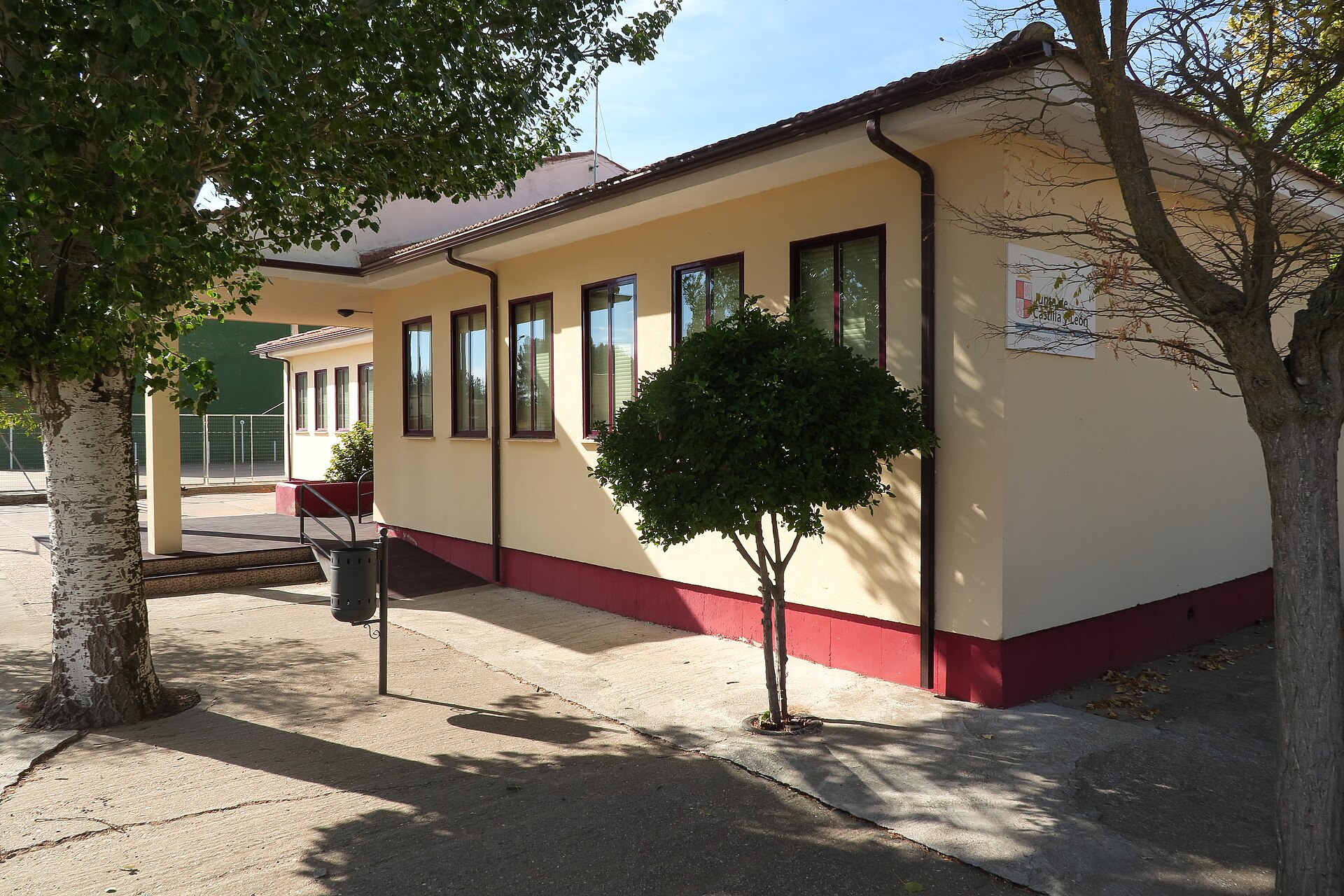 Exterior de un edificio escolar de una planta, con árboles y una pista cercada.