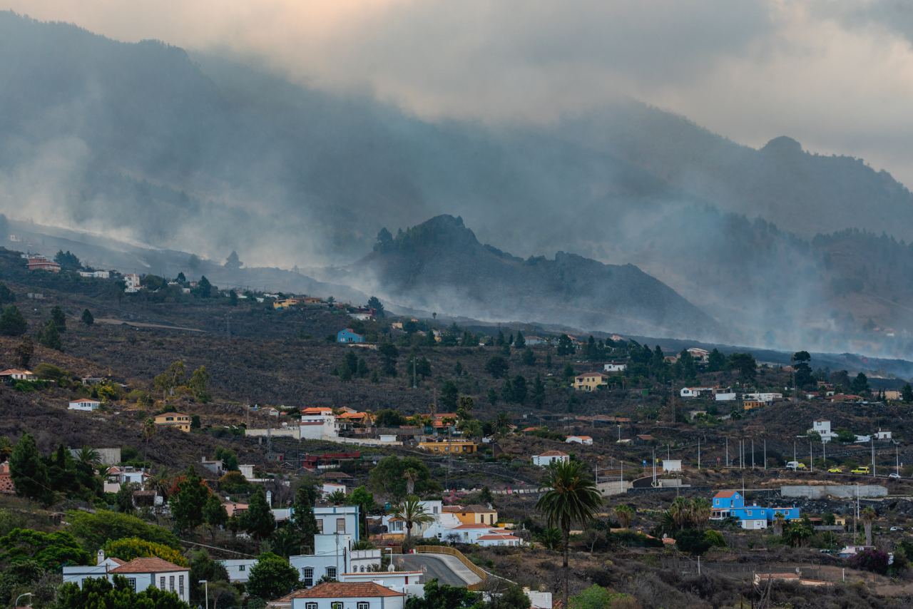 Viviendas y terrenos oscuros se extienden ante un paisaje montañoso cubierto de humo.