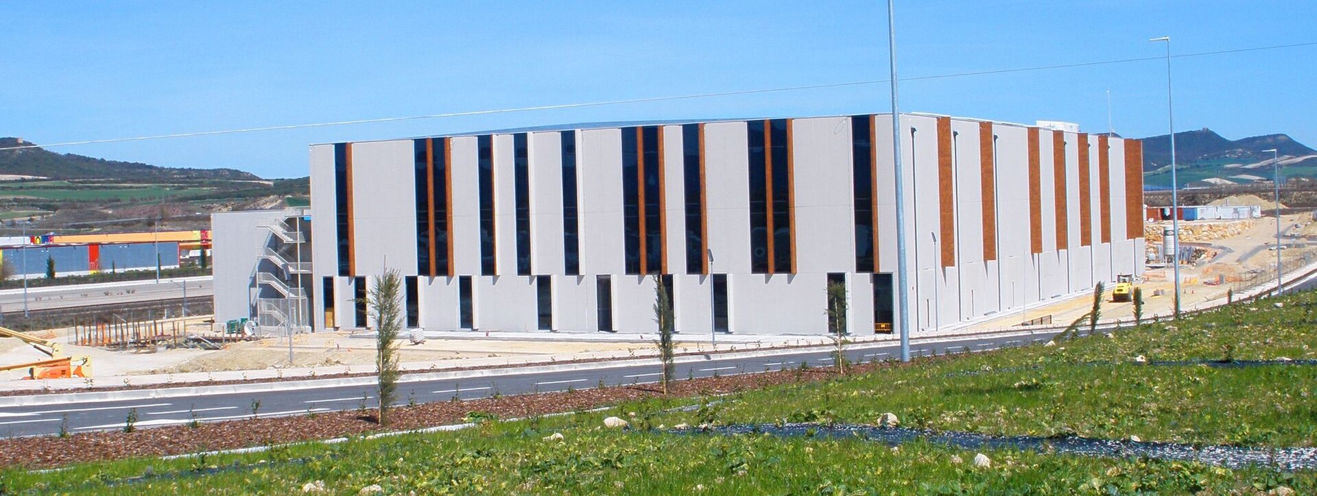 Edificio de gran tamaño con fachada blanca y paneles verticales oscuros y de madera, junto a una carretera y maquinaria de obra.