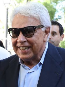 Un hombre de pelo blanco y gafas de sol sonríe, con chaqueta oscura y camisa clara.