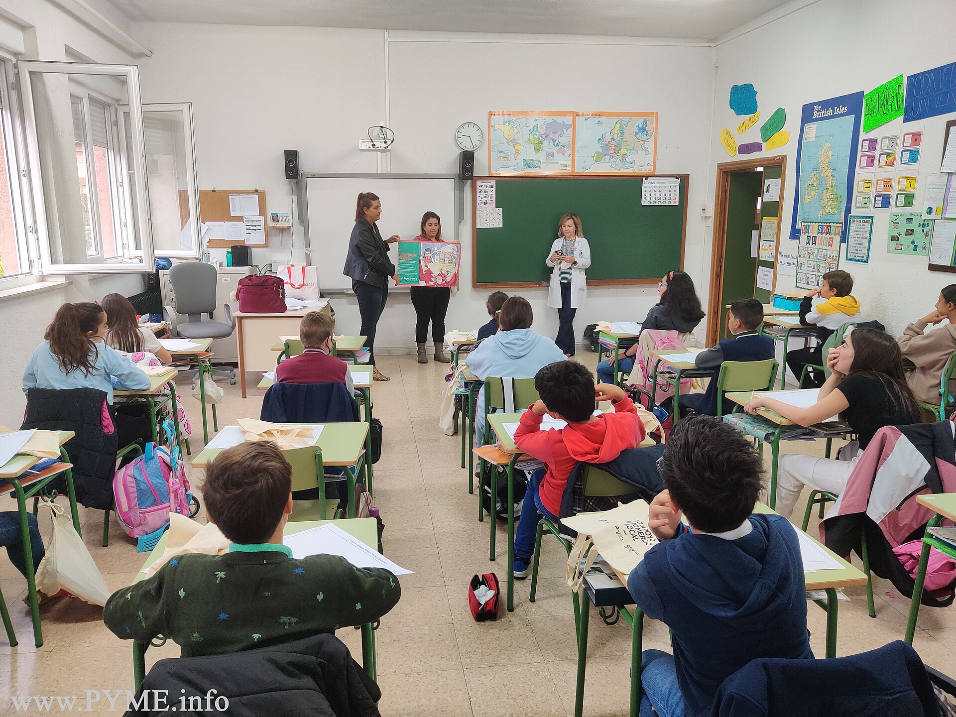 Un grupo de alumnos permanece sentado en un aula mientras varias adultas muestran un cartel.
