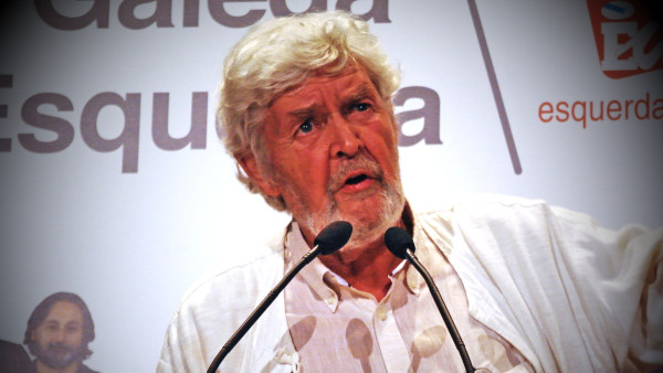 Un hombre de cabello y barba blancos habla ante dos micrófonos.
