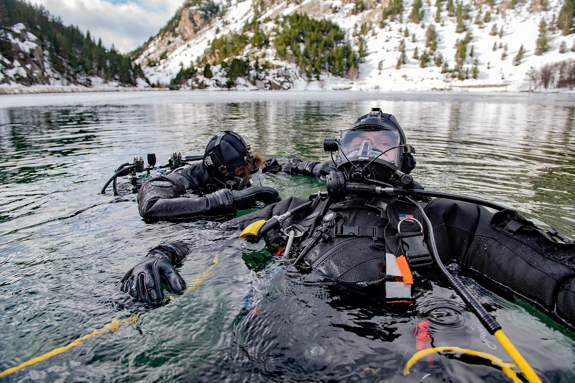 Dos personas con trajes de buceo permanecen en el agua junto a una zona cubierta de nieve.