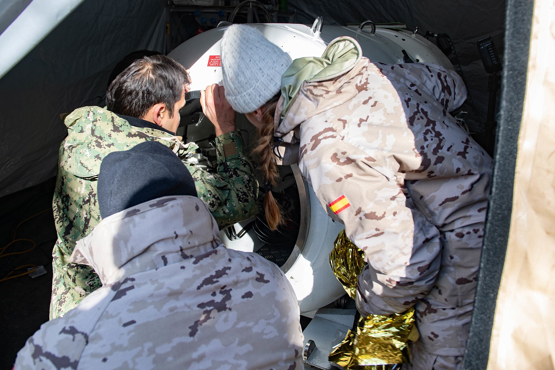 Varias personas con uniformes militares se reúnen junto a una cámara cilíndrica; una de ellas se inclina hacia su abertura.