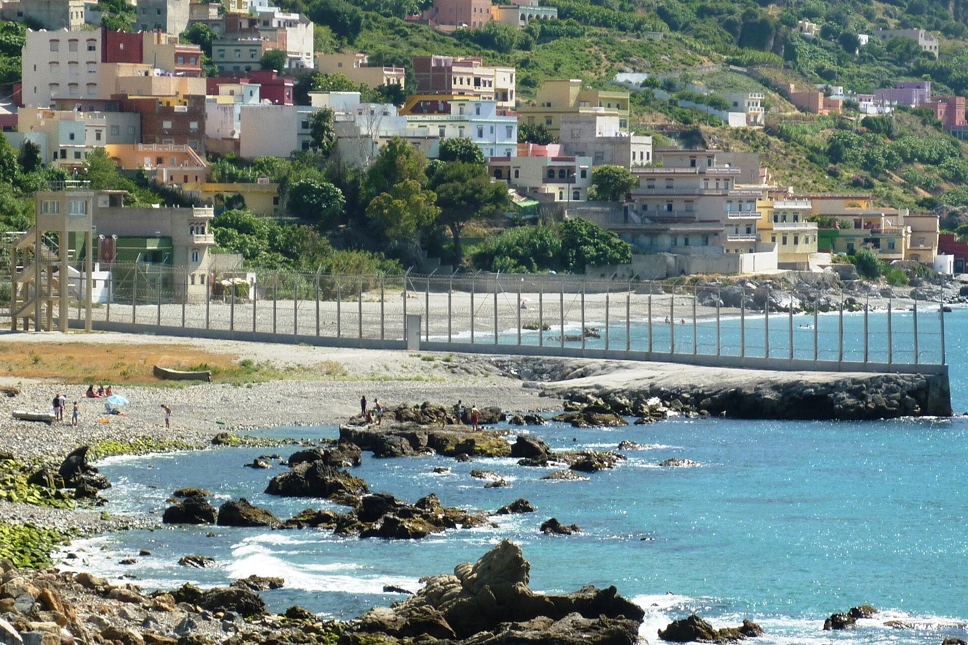 Valla metálica junto a una costa rocosa, con un espigón que se adentra en el mar.