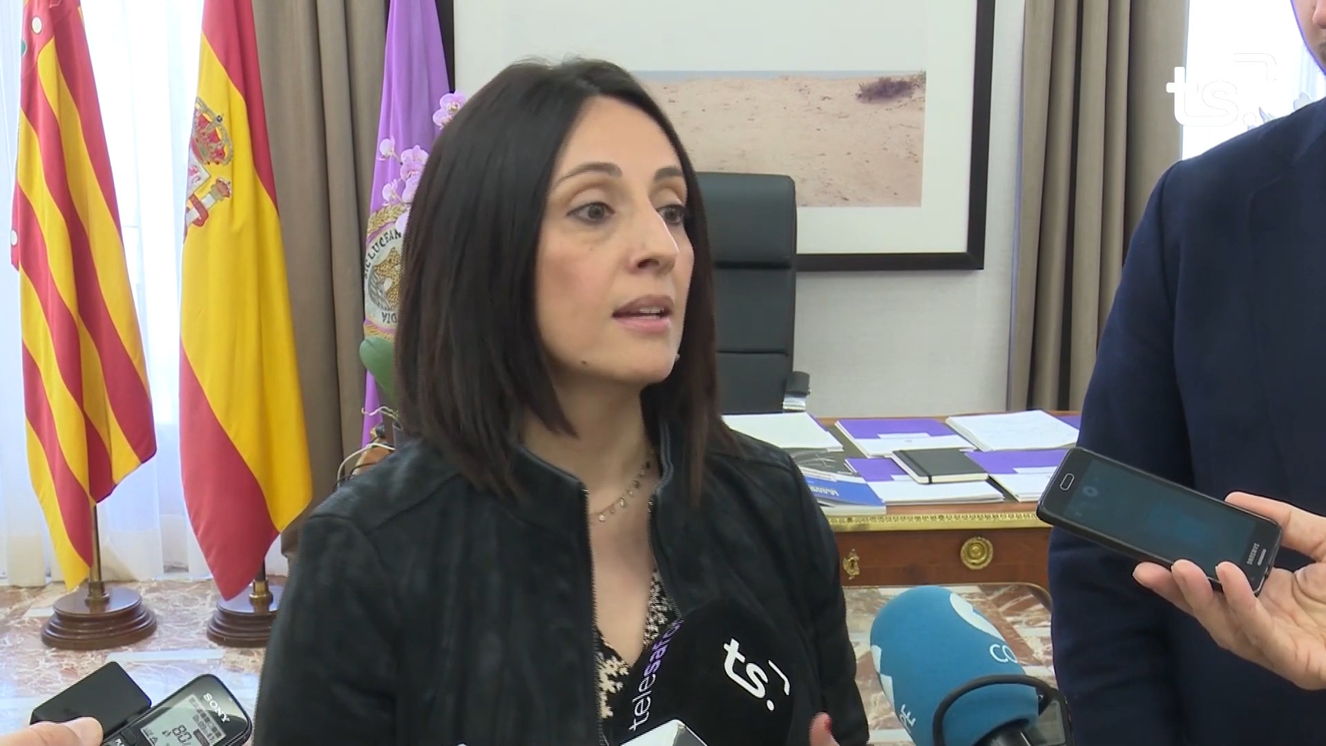 Una mujer atiende a varios micrófonos en una sala con banderas y una mesa de trabajo al fondo.
