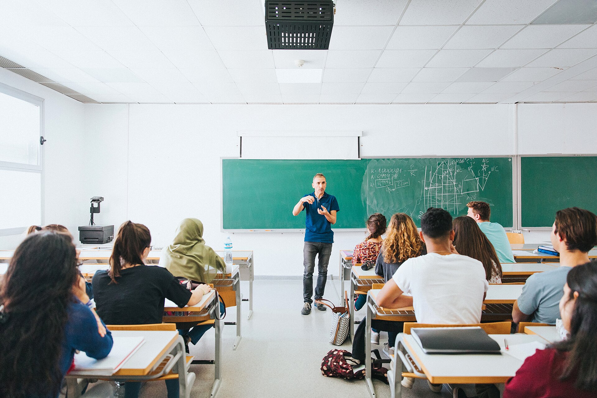 Un docente imparte clase ante un grupo de estudiantes, con fórmulas y gráficos dibujados en la pizarra.