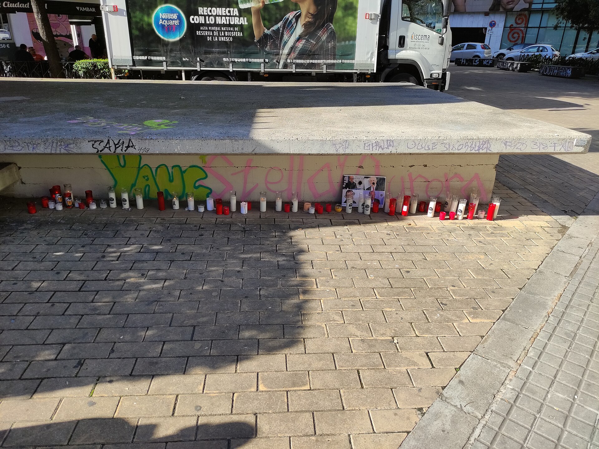 Varias velas están colocadas junto a una pared con pintadas, bajo un camión estacionado.