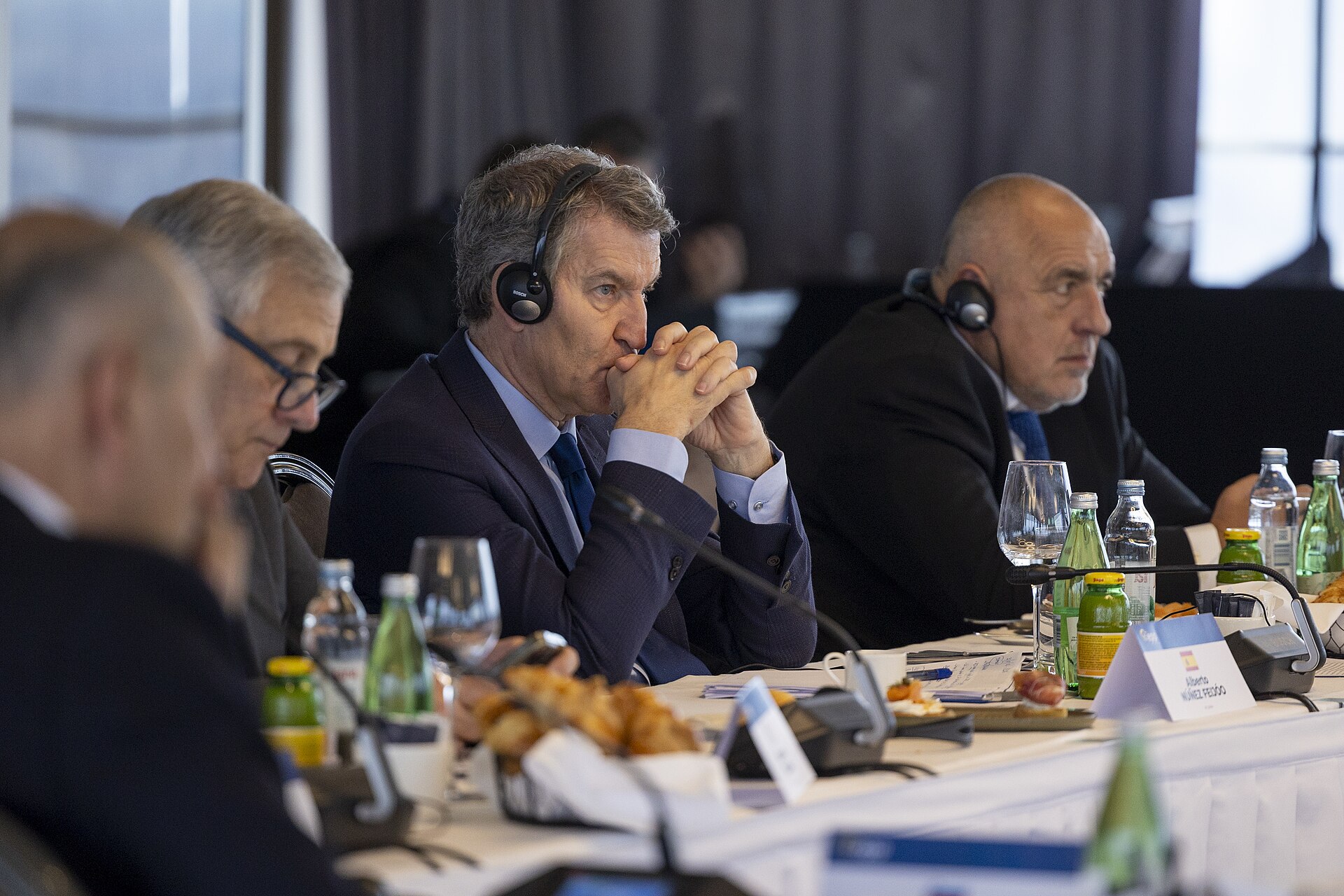 Un hombre con auriculares escucha con las manos juntas en una mesa de reunión, junto a otros asistentes.