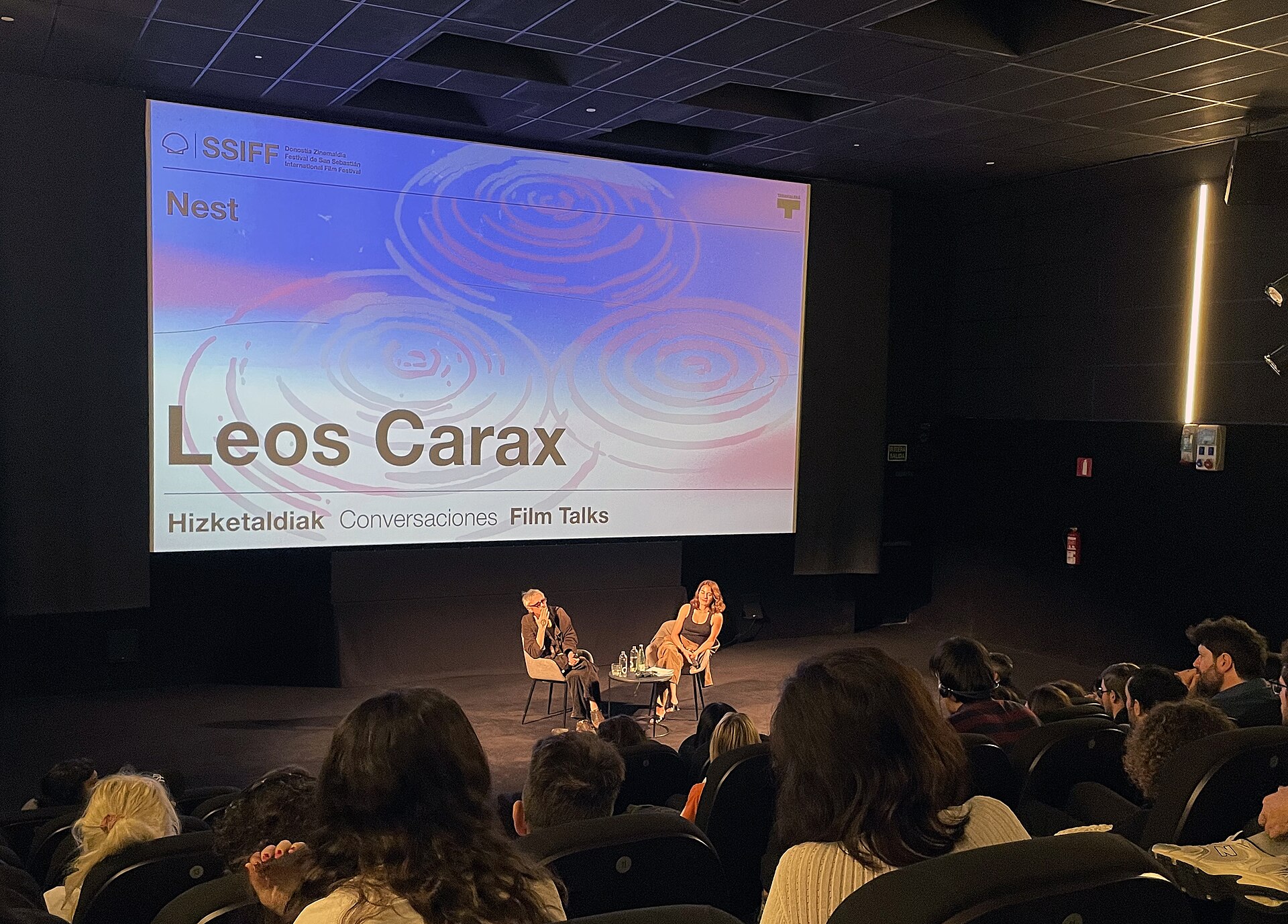 Dos personas conversan en un escenario ante una pantalla que anuncia una charla cinematográfica, mientras el público observa.