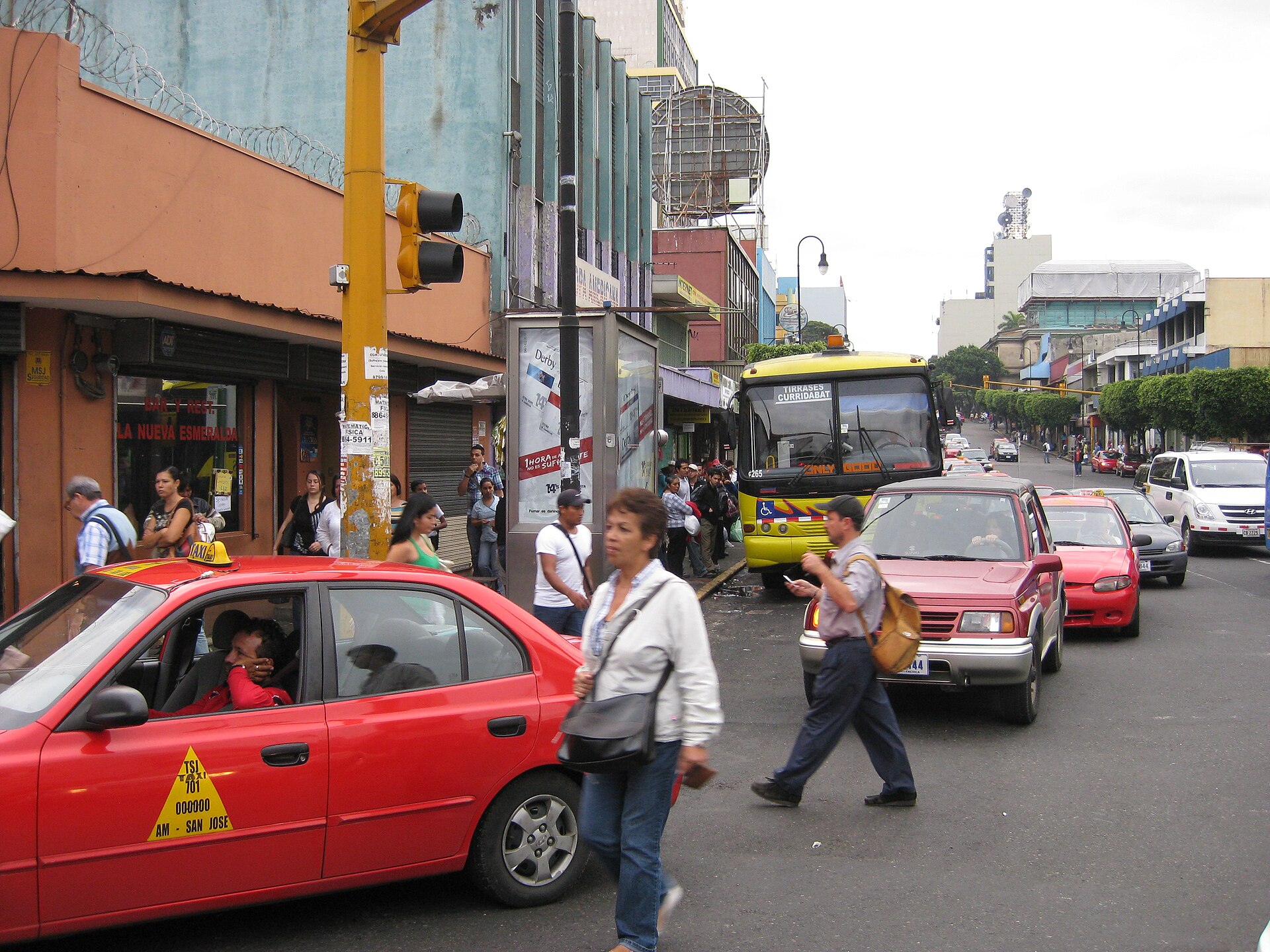 Tráfico de taxis, autobuses y peatones en una calle urbana; uno de los vehículos muestra la inscripción «San José».