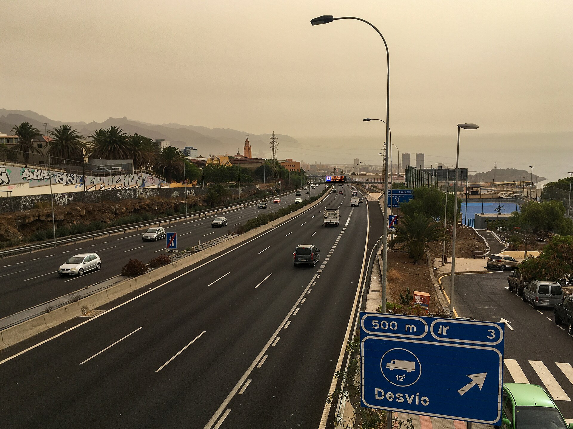 Una autovía con tráfico, palmeras y montañas parcialmente ocultas por una atmósfera amarillenta.