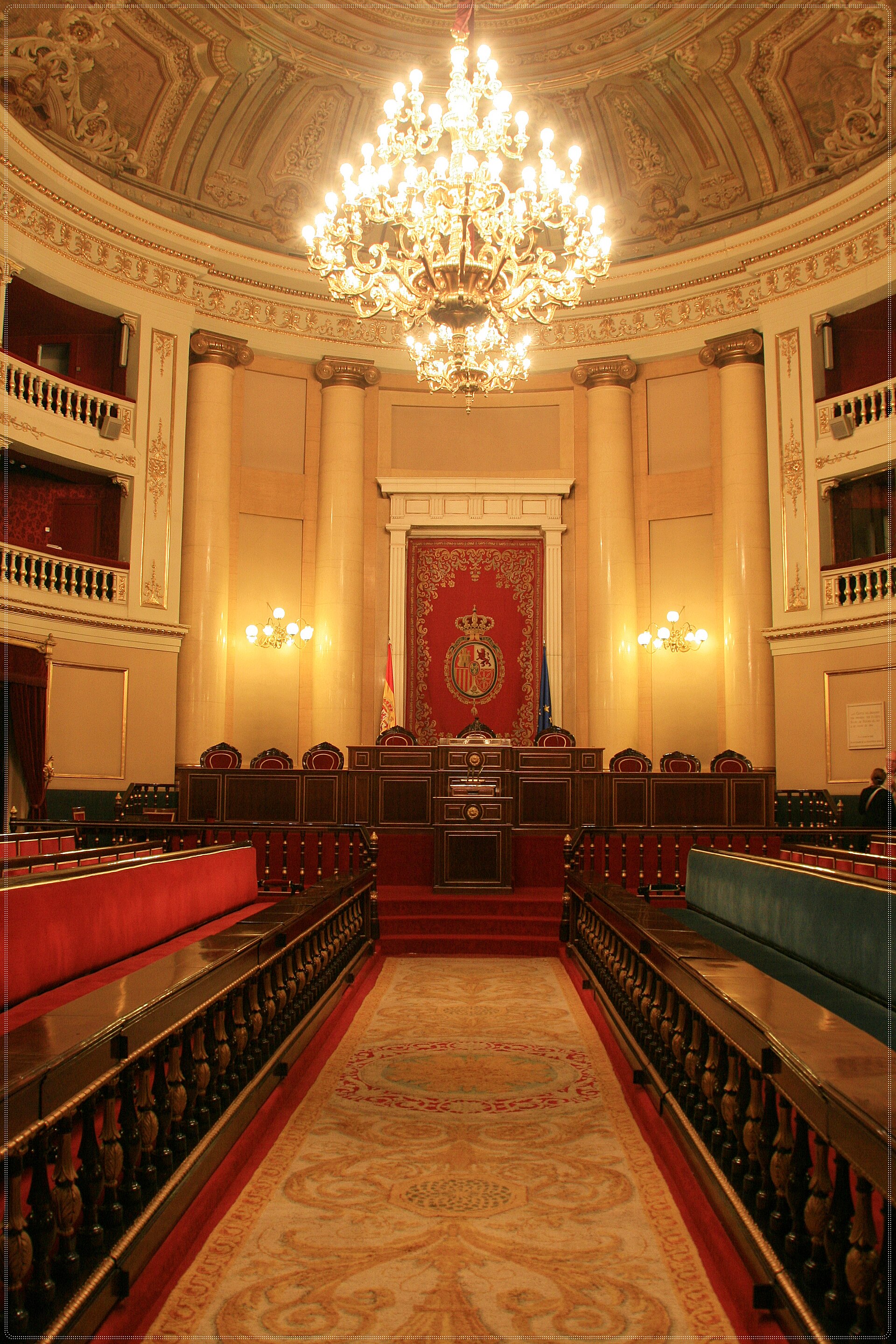 Interior de una sala institucional con bancos, escaños y un escudo bajo un dosel.