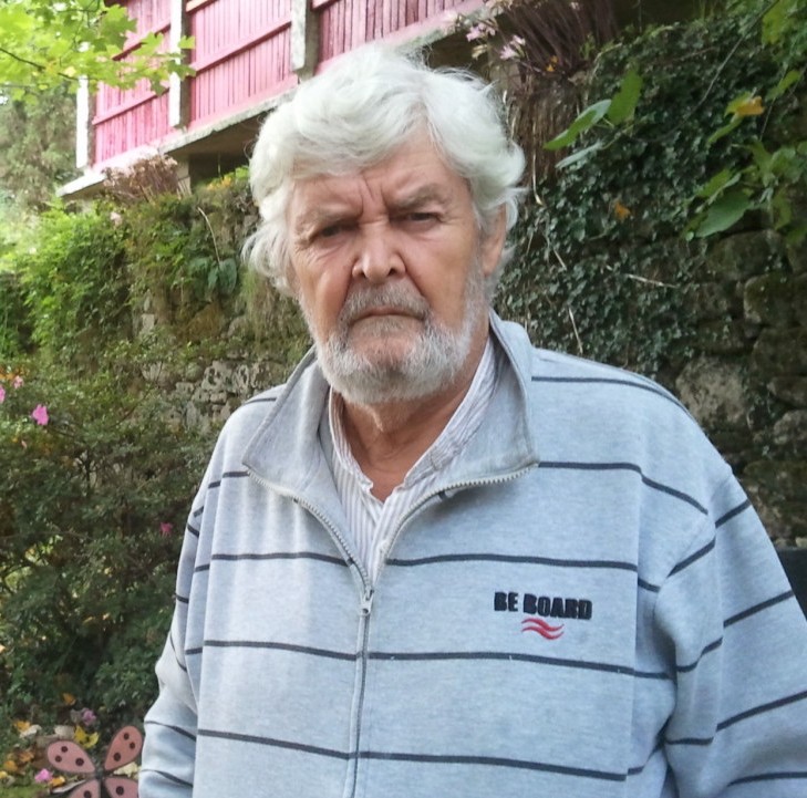 Retrato de un hombre de cabello y barba blancos ante un muro cubierto de vegetación.