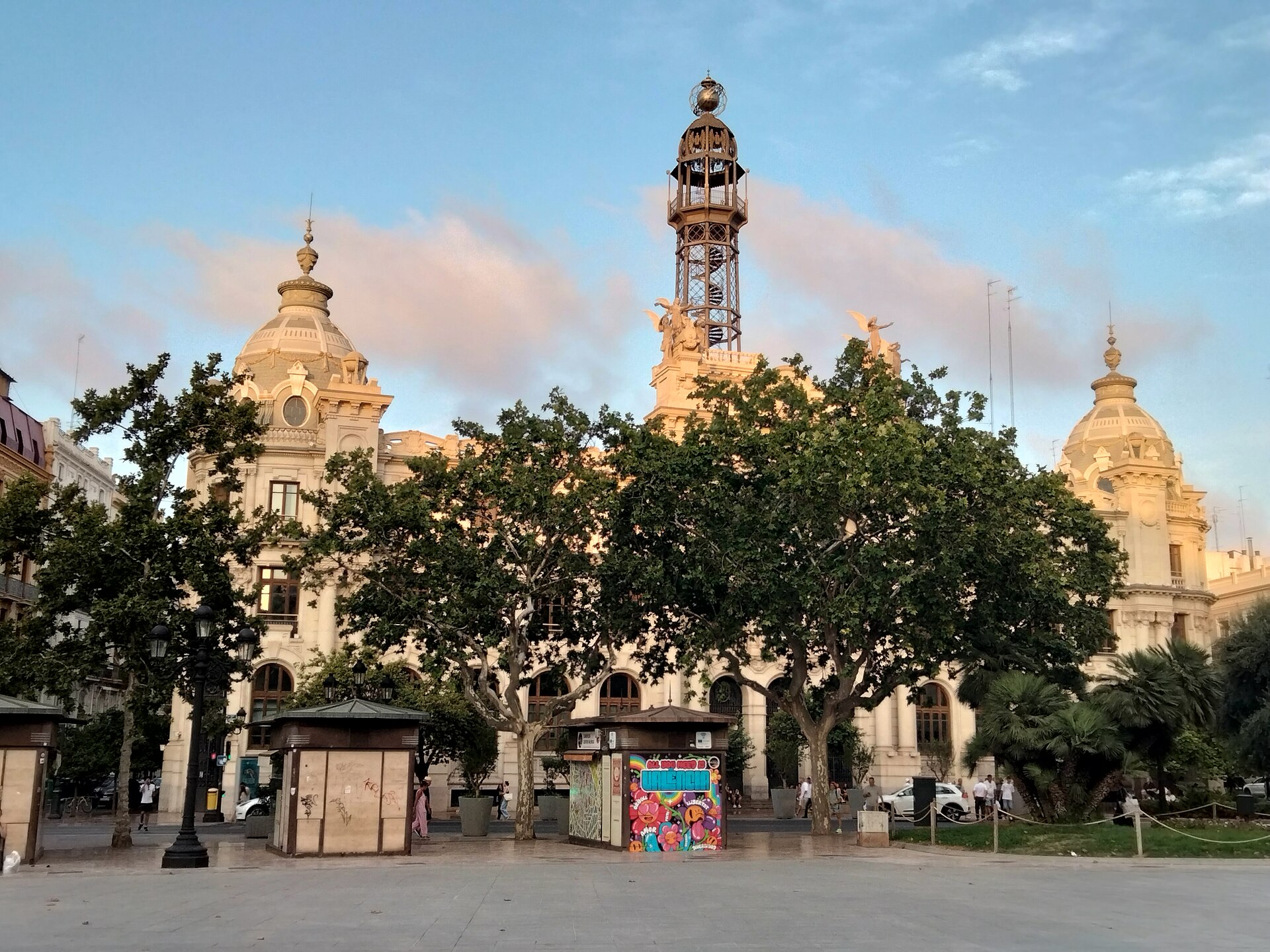 Edificio de piedra clara con una torre central, tras una zona peatonal arbolada.