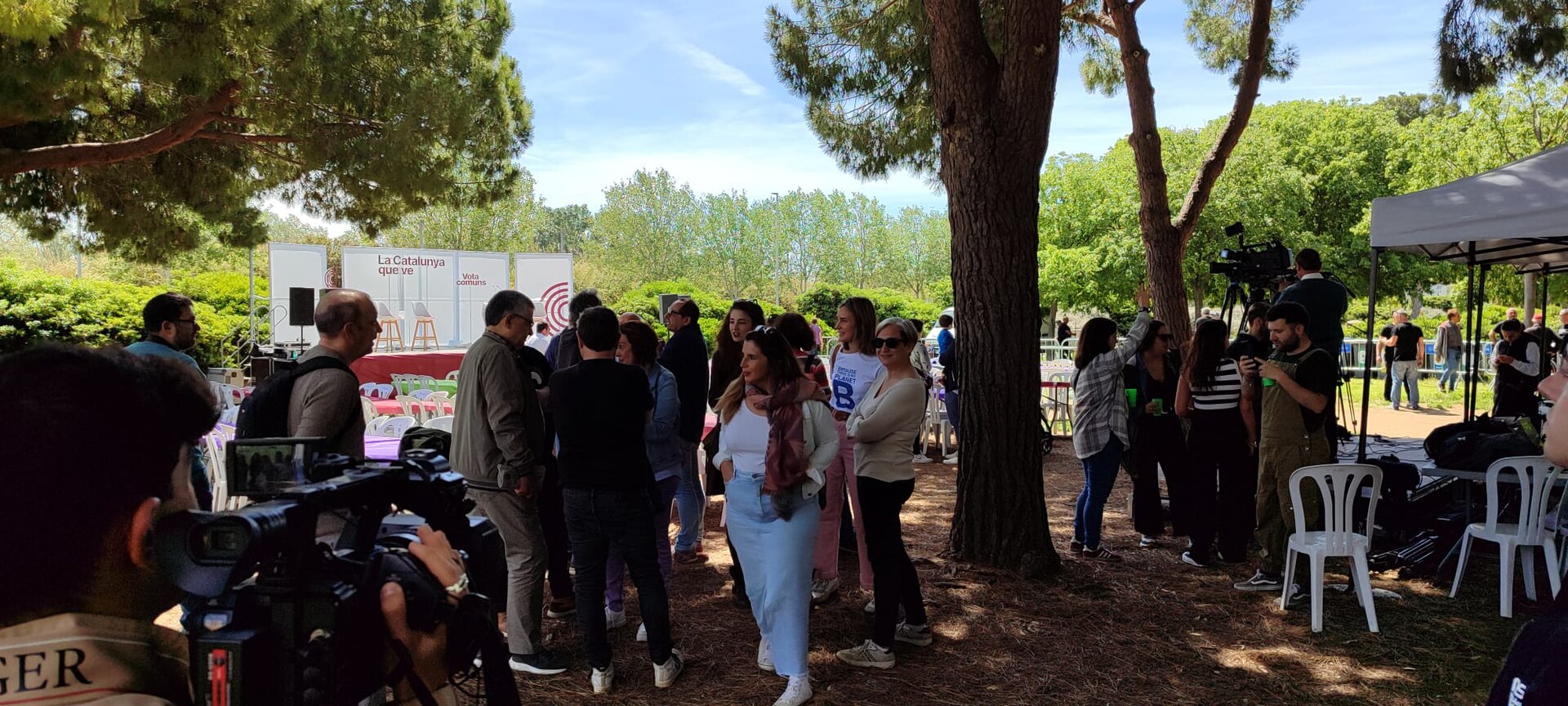 Varias personas conversan ante un escenario con carteles de «La Catalunya que ve» y «Vota Comuns»; se ven cámaras de televisión.