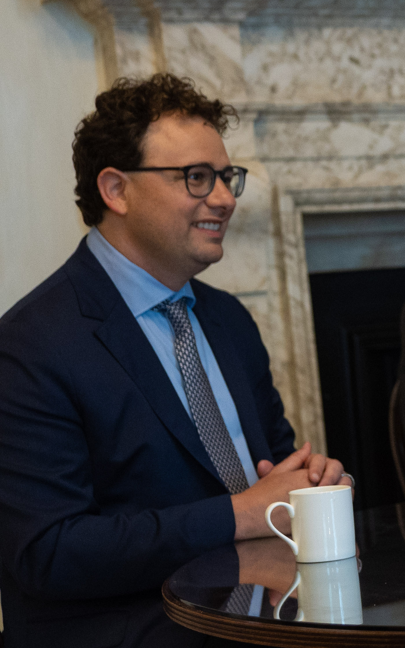 Hombre con gafas y traje sentado junto a una mesa con una taza.