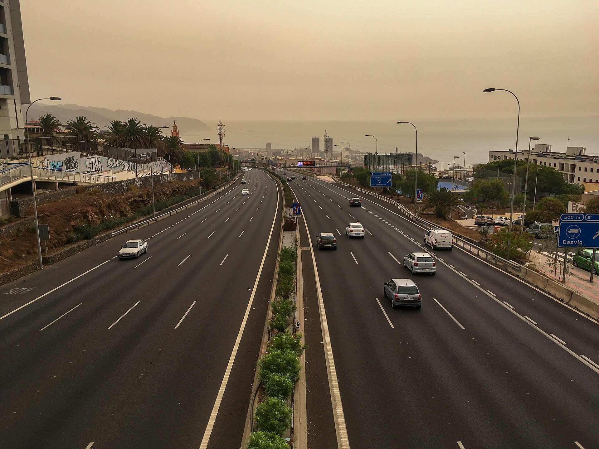 Una autovía de varios carriles bajo un cielo amarillento, con palmeras y edificios al fondo.