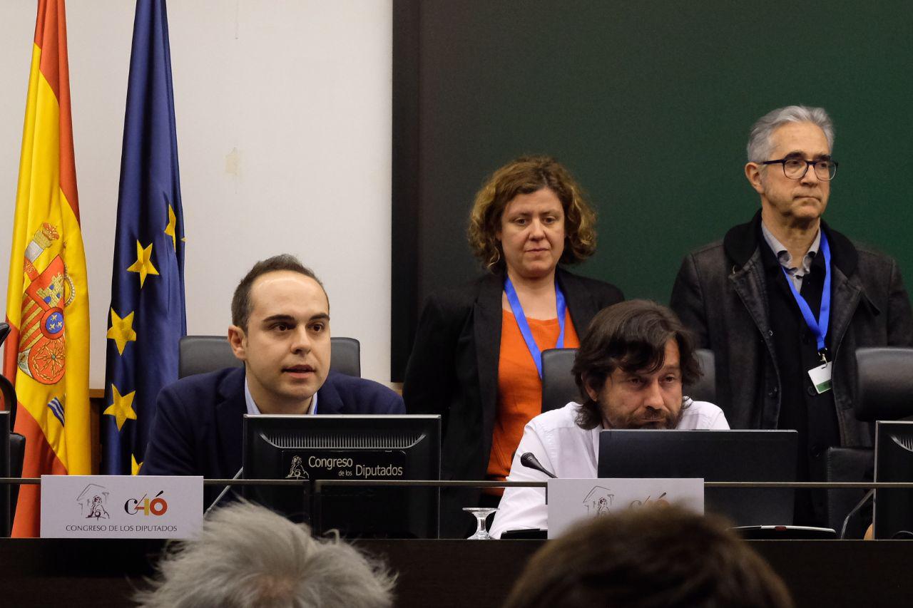 Varias personas sentadas ante pantallas en una sala con las banderas de España y de la Unión Europea; en una pantalla aparece el rótulo «Congreso de los Diputados».
