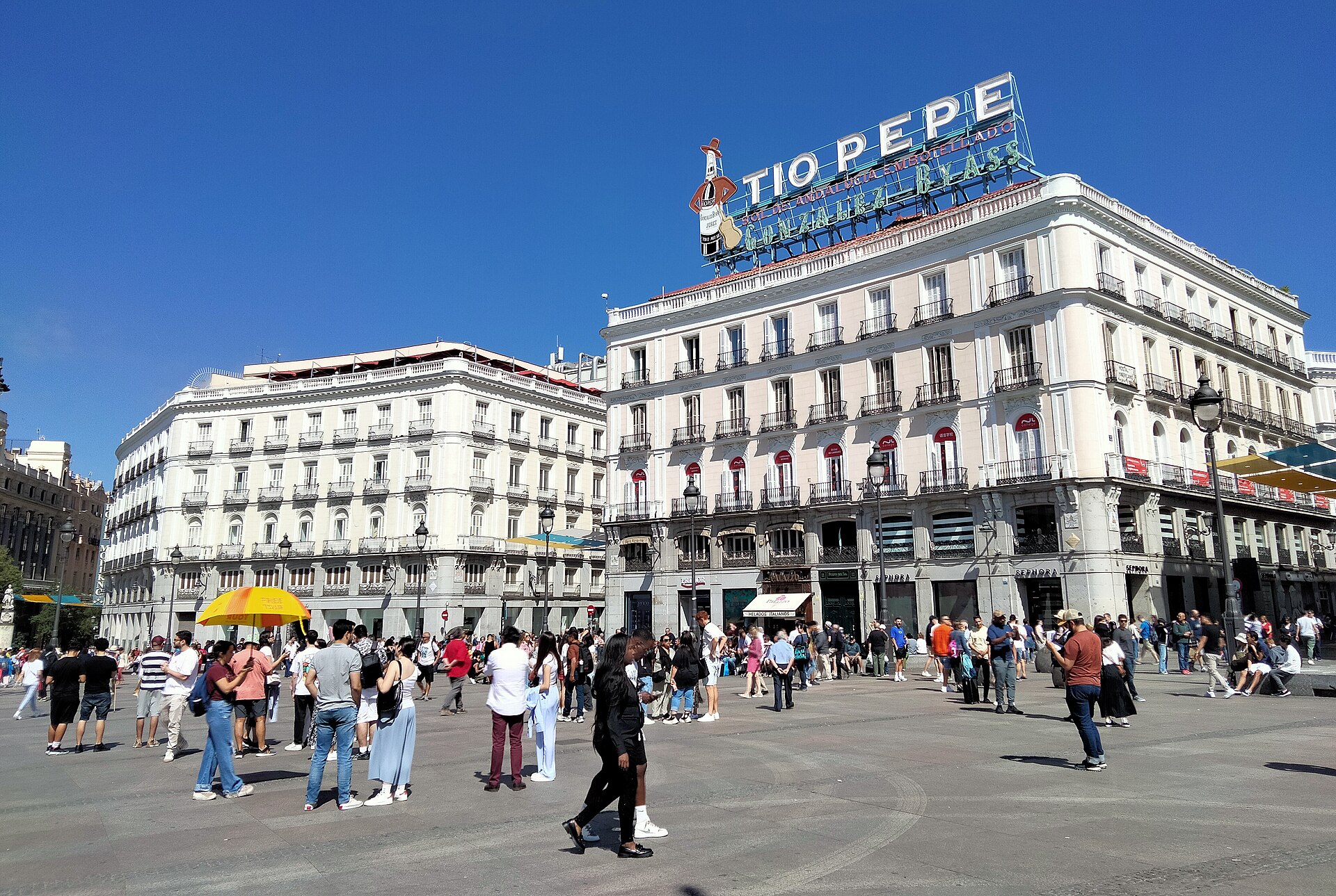 Varias personas caminan por una plaza peatonal frente a edificios con un rótulo luminoso en la azotea.