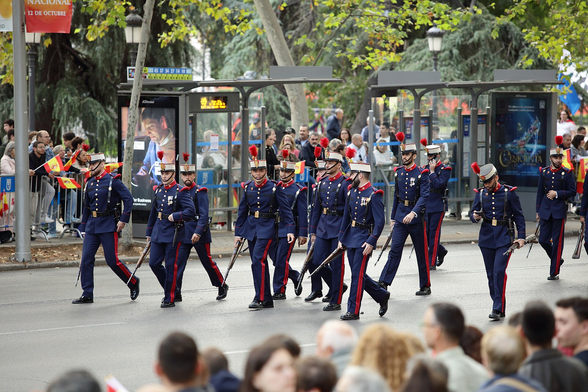 Militares con uniforme azul y penachos rojos desfilan ante un público con banderas españolas.