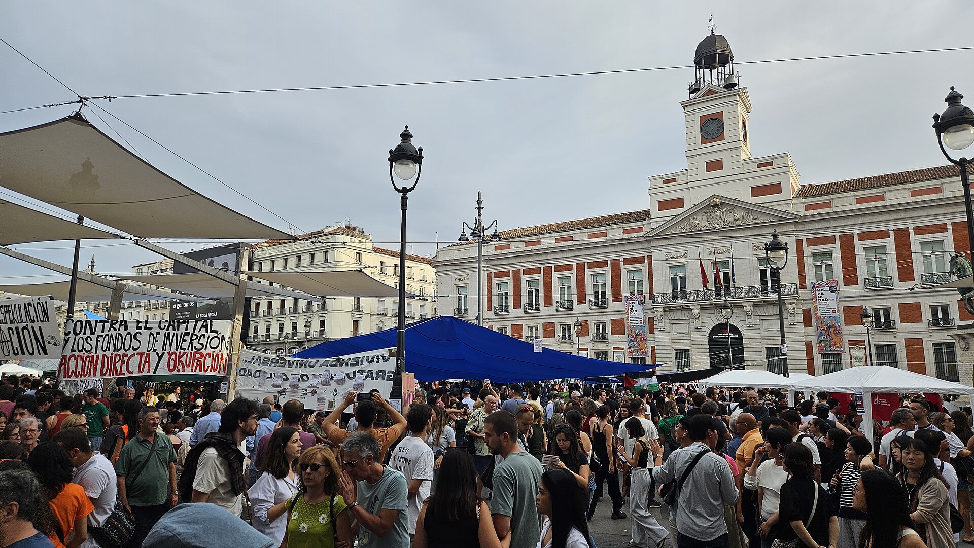 Personas se reúnen entre carpas y pancartas ante un edificio con torre y reloj.