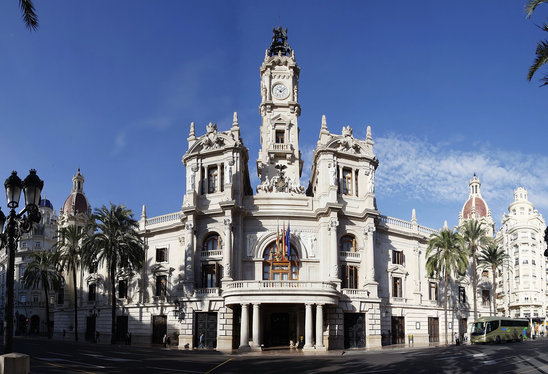 Fachada de un edificio monumental con torre del reloj, palmeras y banderas.