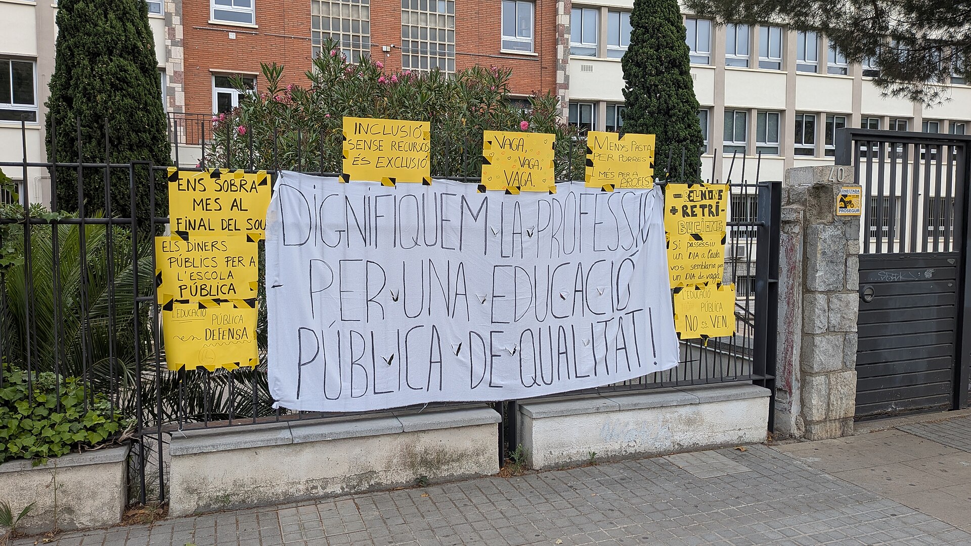Varias pancartas y carteles reivindicativos sobre la educación pública colocados junto a la verja de un edificio.