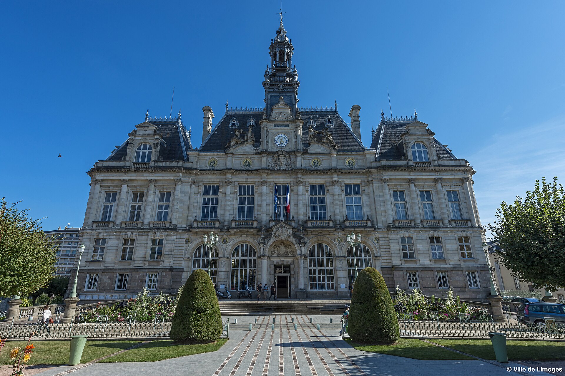 Fachada de un edificio institucional con una torre central, una bandera francesa y jardines delante.