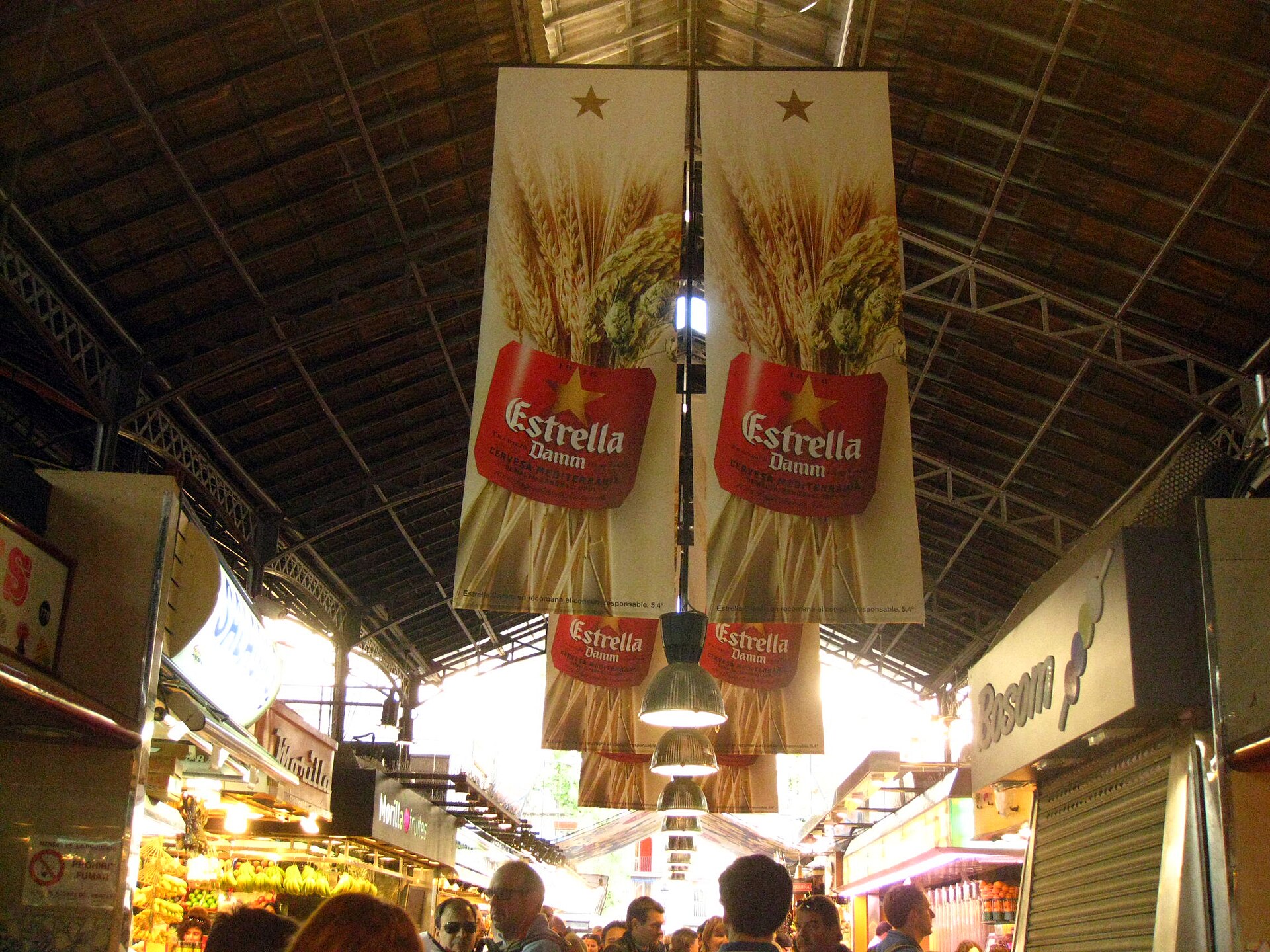 Pasillo interior de un mercado cubierto, con puestos de frutas y verduras y compradores.