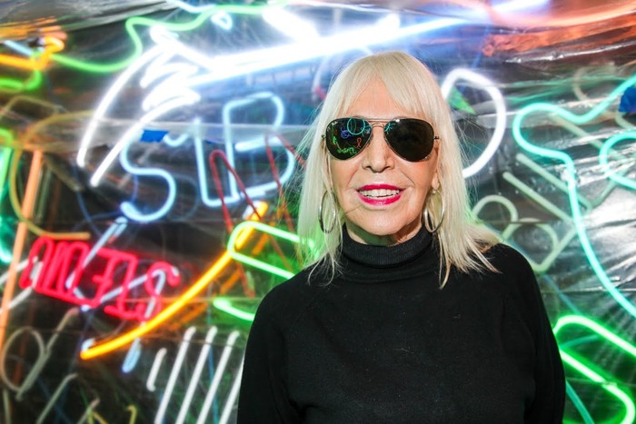 Una mujer con gafas oscuras posa delante de una instalación formada por trazos luminosos de colores.