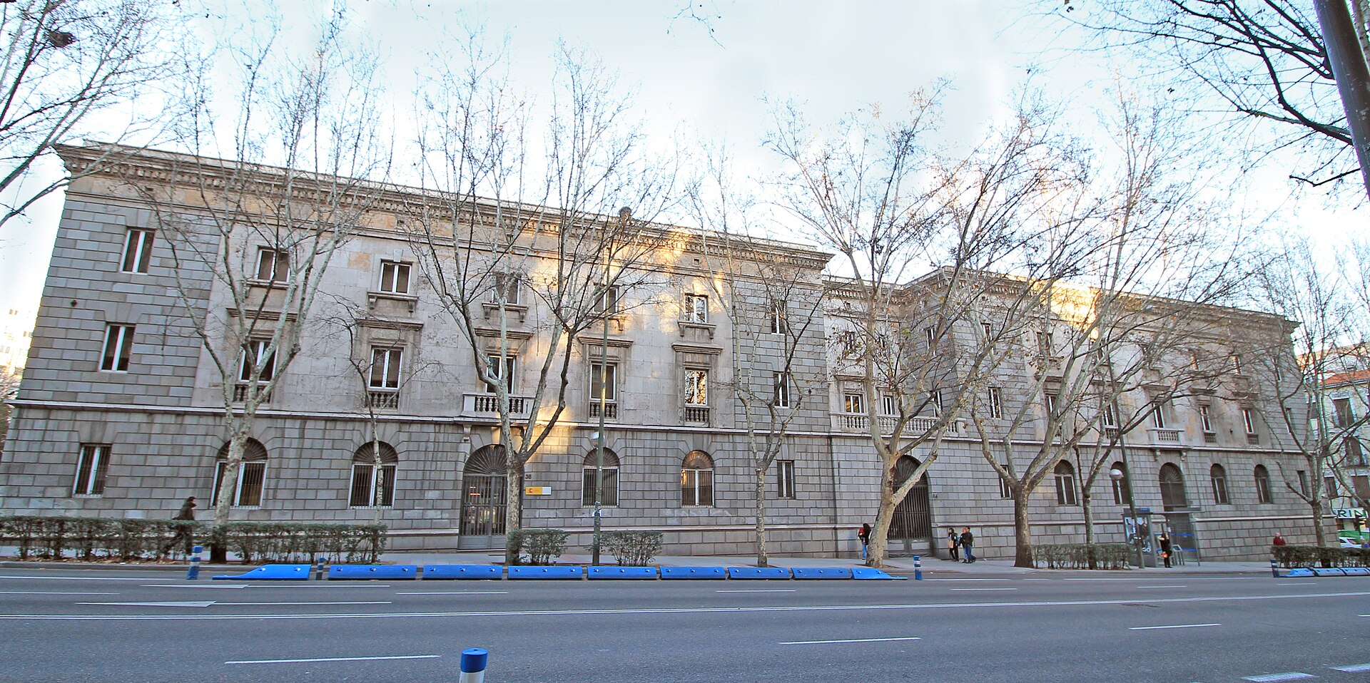 Fachada de un edificio oficial de piedra, parcialmente cubierta por árboles sin hojas.
