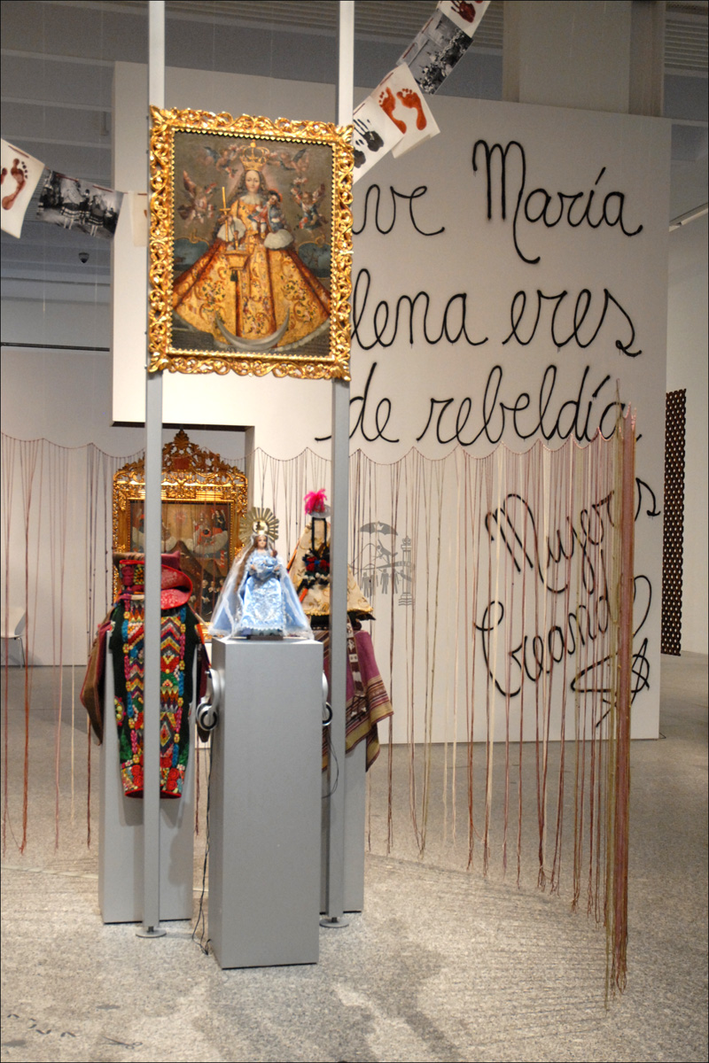 Interior de una sala de exposición con figuras religiosas, textiles y un gran texto mural.